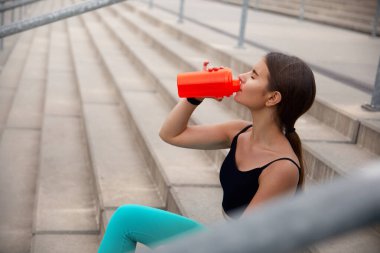 Spor giyim sektöründe aktif genç bir kadın, açık hava basamaklarında mola veriyor, parlak kırmızı bir su şişesinden yudumlayarak, sağlık ve zindeliği vurguluyor. Egzersiz konsepti