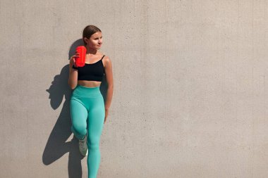 Duvarın önünde eli kalçasında su içen genç bir sporcu. Güneşli bir günde bir kız susuzluğunu giderir. Sıvıyı muhafaza etmek.