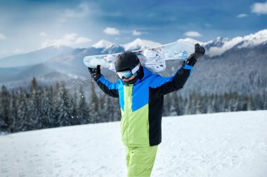 Dağlarda, parlak kış giysileri içinde bir snowboardcu, açık mavi gökyüzü altında, taze kar üzerinde heyecan verici bir inişe hazır bir şekilde sörf tahtasını kaldırır..