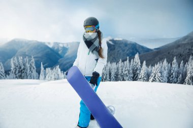 Snowboard 'u olan biri, uzun ağaçlar ve soğuk gökyüzünün altındaki uzak dağlarla çevrili, karlı bir dağda duruyor. Kış sporlarında maceraya hazır.