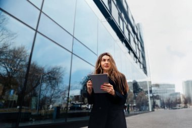 Uzun saçlı genç bir kadın, şehir caddesinde modern binalarla dolu bir tablet tutuyor. Güneşli bir günde çevrenin net yansımalarını sergiliyor..