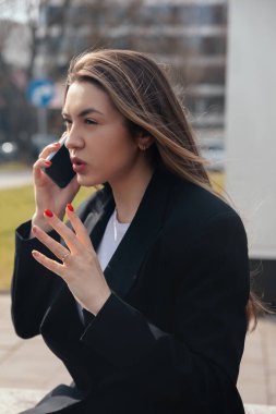 Siyah ceket giyen bir kadın dışarıda telefonuyla tutkuyla iletişim kuruyor. Bir kentin kaldırımında dikilirken elleri duyguları yansıtıyor..