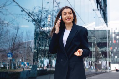 Kendine güvenen bir iş kadını telefonla konuşurken çok hızlı yürür. Etrafı ağaçların ve şehir mimarisinin yansımalarıyla çevrili şık bir kıyafet giyiyor..