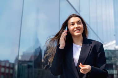 Genç bir kadın dışarıda duruyor, gülümsüyor ve telefonda konuşuyor. Güneş ışığı yakınlardaki cam yapılarda yankılanırken o modern bir kıyafet giyiyor..