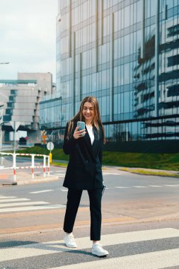 Genç bir kadın, açık bir günde arka planda çağdaş mimari ile telefonunu kontrol ederek güvenli bir şekilde caddenin karşısına geçiyor..