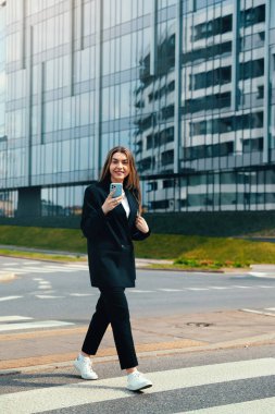 Genç bir kadın şehir merkezinde yaya geçidinde geziyor, elinde akıllı bir telefon, sıradan bir kıyafet giyiyor, özgüvenini göstererek parlak binaların önünde.
