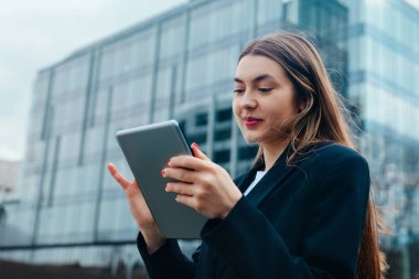 Bir iş kadını kalabalık bir ofis alanında duruyor, koyu renk bir ceket giyerken tabletine odaklanmış. Modern binalar arka planda bulutlu bir gökyüzünün altında..