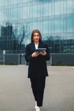Şık siyah bir kıyafet giymiş genç bir kadın, tabletiyle meşgul olan ve sakin bir çevreyle çevrili, çağdaş bir ofis binasının önündeki asfalt bir zeminde sakince yürüyor..