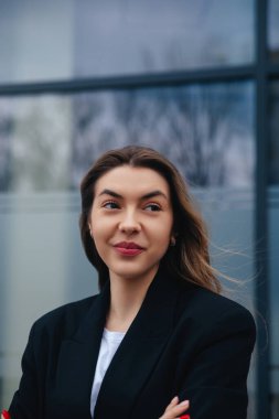 Aydınlık şehir arka planında siyah ceket giymiş gülümseyen genç bir kadın var. Kollarını kavuşturup bu anın tadını çıkarırken kendine güveni tam..