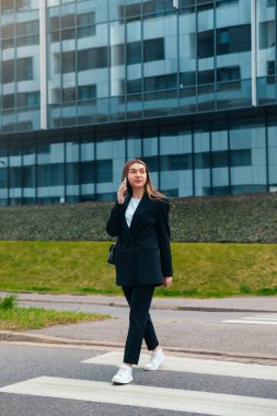 Şık giyinmiş bir kadın kaldırımda yürüyor, kendine güvenen bir tavırla telefonla konuşuyor, şehir ortamında çağdaş bir cam binanın önünden geçiyor..