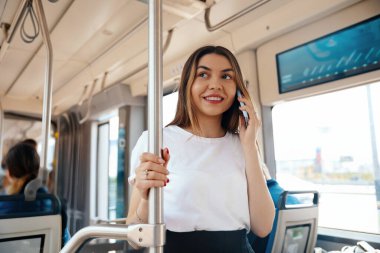 Genç bir kadın halka açık bir otobüste duruyor, kulağına telefon dayayıp gülümsüyor. Güneş ışığı pencerelerden süzülüyor, özelliklerini ve otobüsün içini aydınlatıyor..