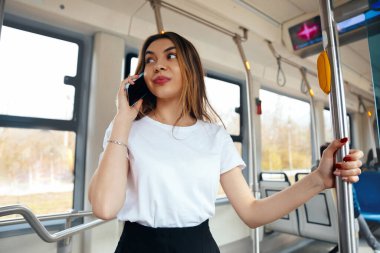 Sıradan giyinmiş bir kadın otobüsün içinde duruyor, bir eliyle direğe tutunarak bir telefon görüşmesi yapıyor, pencerelerden gelen parlak doğal ışıkla çevrili..