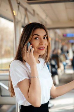 Yoğun bir toplu taşıma aracında telefonla konuşurken gülümseyen bir kadın, pencerelerden süzülen doğal ışık, çevresindeki çeşitli etkinliklerle meşgul olan banliyö sakinleri..
