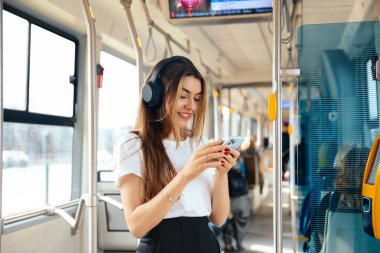 Uzun saçlı genç bir kadın, gündüz saatlerinde toplu taşıma aracında akıllı telefonuna gülümserken kulaklıkla müzik dinlemekten hoşlanıyor..