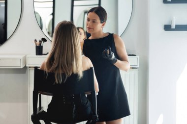 Bir güzellik uzmanı şık bir salonda oturan müşterisine ustalıkla makyaj yapıyor. Atmosfer parlak ışıklandırma ve zarif mobilyalarla dolu, rahatlatıcı bir deneyim yaratıyor..