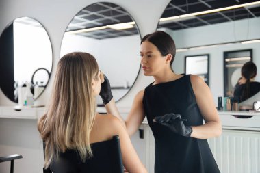 Şık bir güzellik stüdyosunda, makyaj sanatçısı siyah sandalyeye oturan bir müşteriye dikkatlice makyaj malzemesi uygular. Aynalar odaklanmış ifadelerini ve iyi düzenlenmiş alanlarını yansıtır..