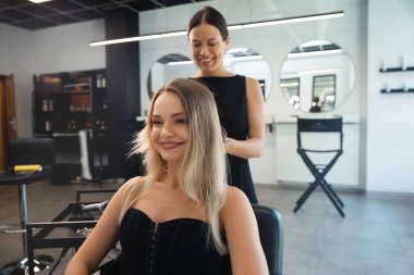 Bir kuaför şık bir kuaförde müşterinin saçı üzerinde dikkatle çalışıyor. Müşteri gülümser, şımartma deneyiminin tadını çıkarır. Doğal ışık boşluğu doldurur..