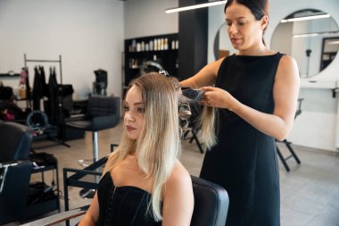 Yetenekli bir stilist modern bir kuaförde uzun saç stili yapar. Müşteri otururken rahat görünüyor, zarif elbisesini ve saç stilini gösteriyor..