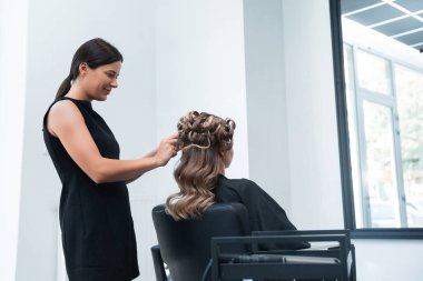 Saç stilisti, geniş pencereli ve parlak doğal ışığı olan şık bir salonda müşterinin saçını kıvırarak rahat ve profesyonel bir atmosfer sunuyor..