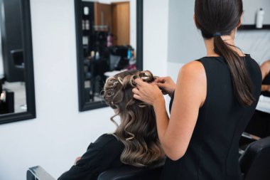 Bir stilist, sakin bir kuaför ortamında müşterisinin saçını ustaca kıvırır. Aynalar profesyonel atmosferi yansıtır ve öğleden sonraları işine odaklanır..