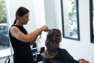 Bir saç stilisti, müşterinin saçlarını, parlak ve çağdaş bir kuaför salonunda, kişisel dönüşüm ve özen anı yakalayarak, güzel kıvırcık saçlara dönüştürür..