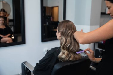 Stilist, müşterinin saçını ustalıkla kıvırıyor. Geniş aynalı çağdaş bir salon ortamında zarif dalgalar yaratıyor..