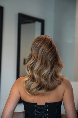 Bir kadın şık bir bluz giyerken güzel dalgalı saçlarını sergiliyor. Sahne modern bir banyoda, parlak aynalar ve yumuşak ışıklandırmayla çekilmiştir..