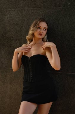 Bir kadın, koyu taştan bir duvarın yanında kendine güvenerek poz veriyor. Moda tercihini siyah elbiseyle sergiliyor. Zarif duruşu zarafetinin altını çiziyor..