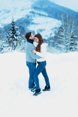 İki insan, arka planda dağlar ve ağaçlar olan sakin bir kış manzarasıyla çevrili karda neşeli bir anı kucaklaşır ve paylaşır..