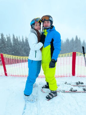 Bir çift renkli kayak kıyafetleri içinde bir kayak merkezinde mutlu mesut dururlar. Karlı ağaçlar ve bulutlu gökyüzü ile çevrili yamaçlara çıkmaya hazırlar..