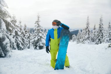 Kar kaplı bir yamaçta parlak kış kıyafetleri giymiş bir snowboardcu duruyor. Arka planda bulutlu bir gökyüzünün altında, kışın güzelliğini gözler önüne seren uzun ve yeşil ağaçlar var.