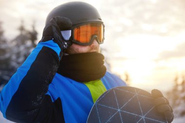 Snowboardcu, ağaçlarla çevrili karlı bir yamaçta ve batan güneşle aydınlatılmış güzel bir gökyüzünde dururken vites ayarlar. Kış sporu