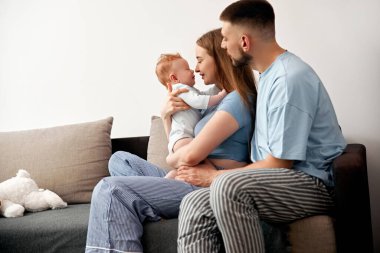 Parlak yaşam alanındaki rahat bir kanepede neşeli bir etkileşim içinde olan ebeveynler ve bebeklerle aile anlarını sevmek sıcaklığı ve bağlantıyı ön plana çıkarıyor.