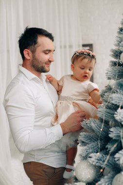 Bir adam ve küçük kızı neşeli bir odada Noel ağacını süslerken tatlı bir anı paylaşıyorlar..