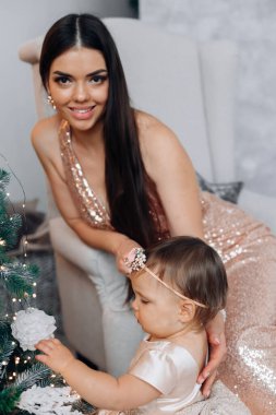 Zarif payetli bir elbise giymiş bir anne kameraya gülümserken küçük kızı evdeki güzel süslenmiş bir Noel ağacında süs eşyalarına uzanır..