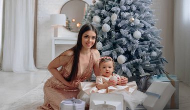 Neşeli bir sahnede bir anne bebeğiyle birlikte güzel paketlenmiş hediyelerle çevrili bir Noel ağacının yanında bir aile kutlaması anını yakalıyor..