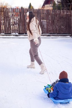 Mavi plastik bir kızağın üzerinde oturan küçük bir çocuğu çekerken bir kadın karda yürüyor. Kadın ve çocuk kahverengi ahşap bir çitin yanında bir avludalar..