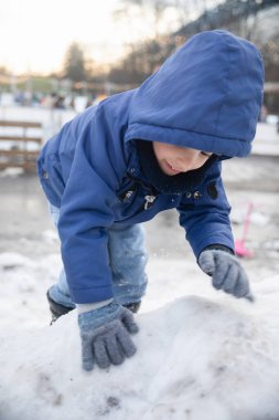 Eldivenli elleri olan küçük bir çocuk parkta kardan bir şey yapıyor. Çocuk mavi bir ceket giyiyor ve zemin karla kaplı..