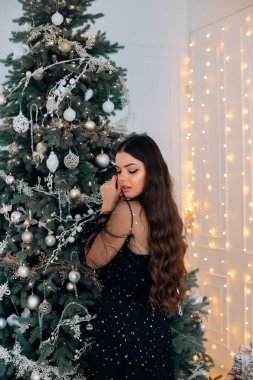 Genç bir kadın güzel süslenmiş bir Noel ağacının yanında, sıcacık aydınlatılmış bir odada zarif bir şekilde duruyor. Sahne, tatil sezonunun neşeli ruhunu yakalıyor..