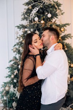 Bir erkek ve bir kadın zarif bir Noel ağacının önünde sevgi dolu bir şekilde kucaklaşıyorlar. Işıklar ve süslemelerle süslenip tatil sezonu için sıcak ve şenlikli bir ortam yaratıyorlar..