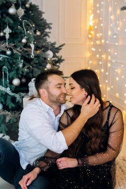 Bir çift, tatil süslemeleriyle dolu bir odada yan yana oturuyor. Güzel dekore edilmiş bir Noel ağacı ve yumuşak ışıklarla çevrelenmiş bir şekilde hassas bir anı paylaşıyorlar..