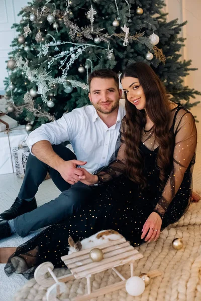 Bir adam ve bir kadın güzel süslenmiş bir Noel ağacının yanında yumuşak bir halının üzerinde birlikte oturuyorlar. Aynı sevgi dolu bakışları paylaşıyorlar, şık bir tatil kıyafeti giyiyorlar, neşeli ve samimi bir atmosfer yaratıyorlar..