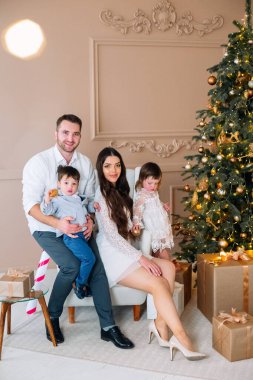 Mutlu bir aile, güzel dekore edilmiş bir Noel ağacıyla şık bir odada, hediyelerle çevrili bir şekilde poz verir. Tatil sezonunda bir sevinç ve kutlama anı paylaşıyorlar..