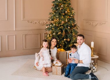 Bir aile, süslenmiş bir Noel ağacının yanında altın süsler ve ışıklarla bir araya gelir. Ebeveynleri yanlarında otururken iki çocuk gülümsüyor ve sıcak bir tatil ortamı yaratıyorlar..