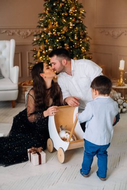 Bir çift, tahta bir oyuncak arabasının yanında diz çökmüş meraklı bir çocuğun içindekileri keşfetmesini izlerken sevgi dolu bir öpücüğü paylaşıyor. Samimi oda şenlikli ışıklar ve bir Noel ağacı ile süslenmiş..