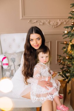 Neşeli bir anne, genç kızıyla birlikte güzel dekore edilmiş bir Noel ağacının yanında oturur. Bayram ışıkları hafifçe parıldarken oda sıcaklığı yayıyor..