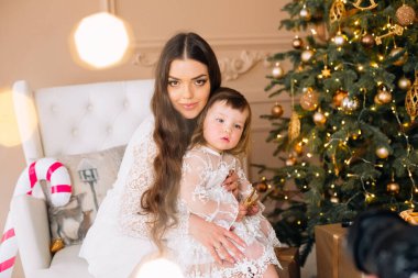 Bir kadın, Noel ışıkları ve altın süslemelerle çevrili rahat bir sandalyede oturup, sıcak bir tatil ortamı yaratarak, küçük çocuğunu kucaklar..