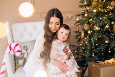 Bir anne, genç kızını çok güzel dekore edilmiş bir Noel ağacının önünde otururken kucağına alır. Işıklar ve süslemeler, tatil ruhuyla dolu rahat bir oturma odasında parlar..