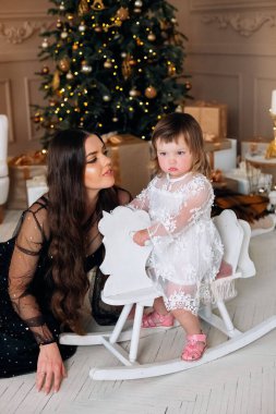 Beyaz elbiseli genç bir kız sallanan bir atın üzerinde otururken annesi yakınına eğiliyor ve güzel dekore edilmiş bir Noel ağacıyla birlikte güzel bir tatil ortamının tadını çıkarıyor..