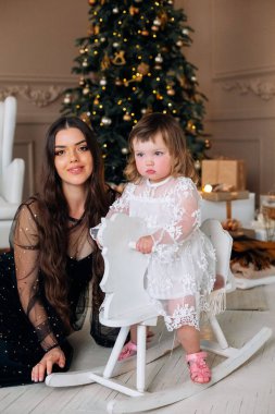 Bir anne ve küçük kızı, Noel ağacıyla süslenmiş bir festival odasında neşe içinde poz veriyorlar. Küçük kız beyaz dantelli bir elbise içinde sallanan bir atın yanında duruyor..